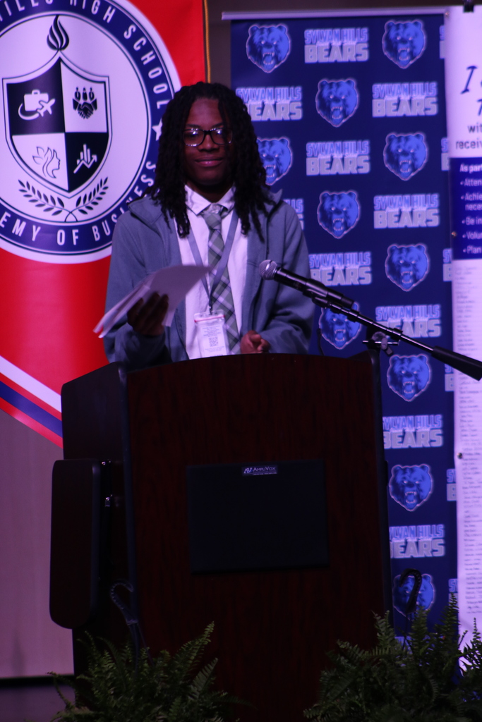 A presenter stands behind a podium with plants in front, speaking in front of a Sylvan Hills Bears backdrop and an Academy of Business banner.