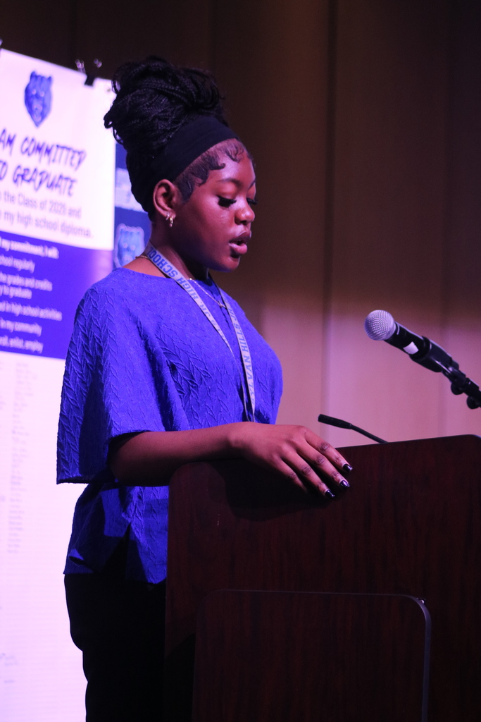 A presenter wearing a blue textured garment and a Sylvan Hills High School lanyard speaks at a podium. Behind them is a large “I Am Committed to Graduate” banner lit with purple stage lighting.