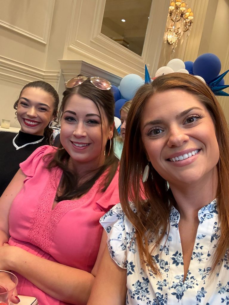 Three people sit together at a table inside a decorated hall with cream walls, ornate molding, and a chandelier. A display of blue and white balloons is arranged behind them as they smile toward the camera.