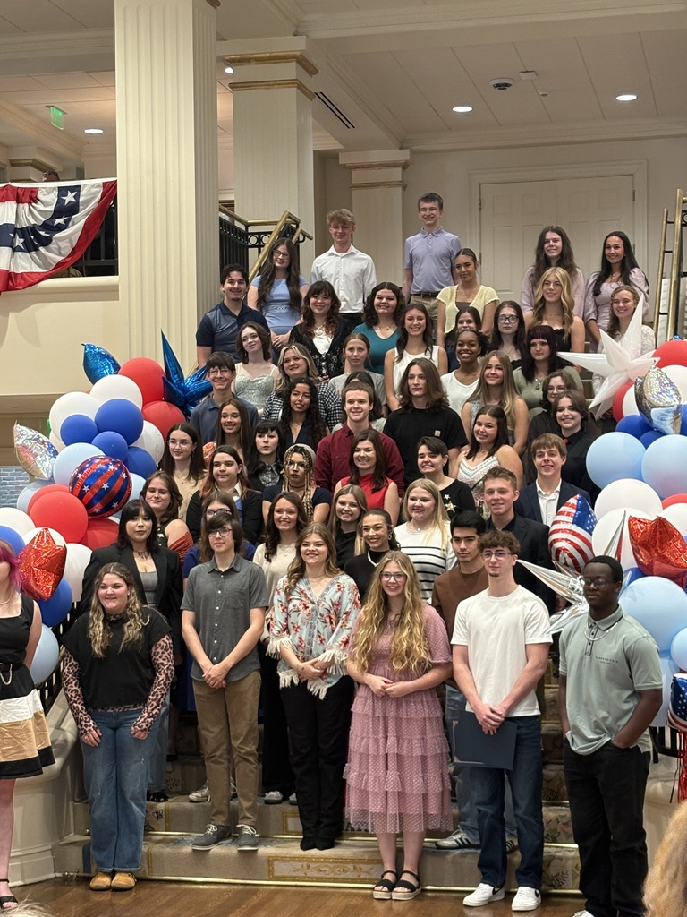 A large group poses on a staircase inside a hall decorated with red, white, and blue balloons and star‑shaped accents. People stand and sit in several rows for a celebratory group photo.