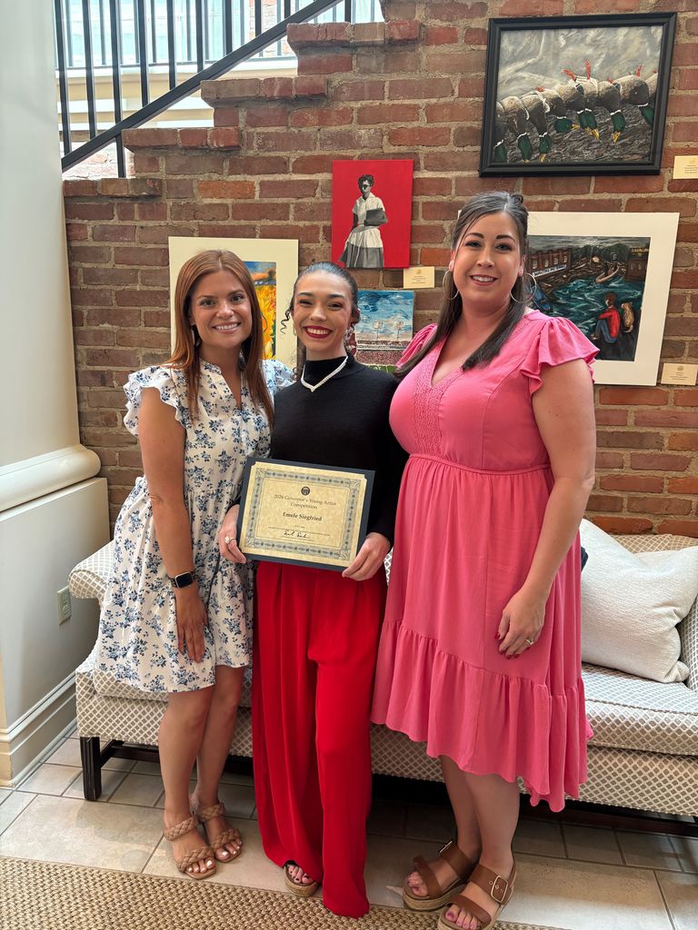 Three people stand in front of a brick wall displaying framed artwork. The student in the center holds a certificate reading “2023 Covington’s Young Artist Emelie Siegfried,” and all three smile toward the camera.