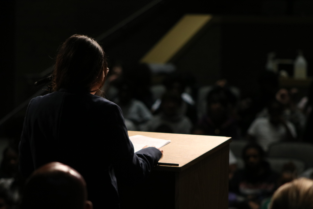 A person stands at a podium holding a sheet of paper while speaking to an audience in a dimly lit auditorium. The audience is visible but out of focus.