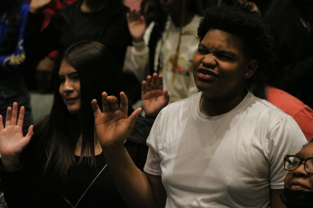 A group of people raise their right hands during a pledge. Two individuals in the foreground, one in a white shirt and one in black, have their hands raised with open palms. Others in the background do the same.