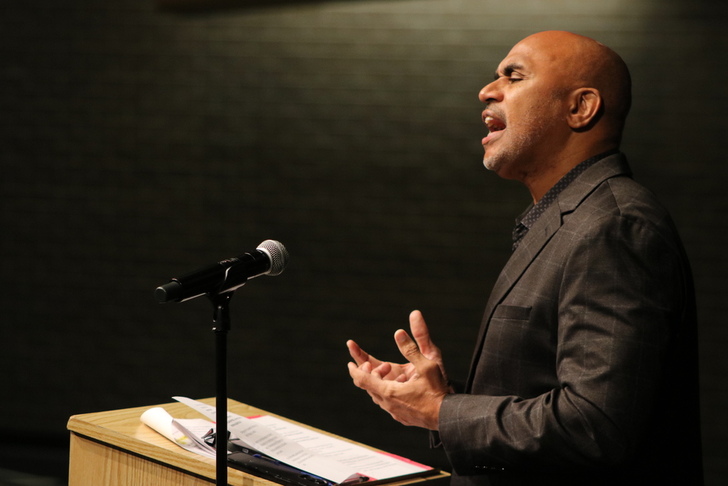 A person in a dark suit stands at a wooden podium speaking into a microphone. Papers and a pen rest on the podium, and the background is dark.