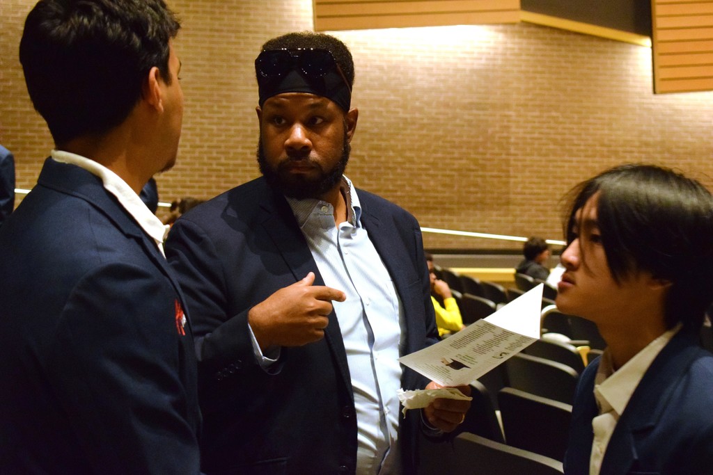 Three people stand together in an auditorium aisle, talking. All are dressed in dark blazers and light shirts. One person in the center holds a printed program. Rows of seats and brick walls are visible behind them.