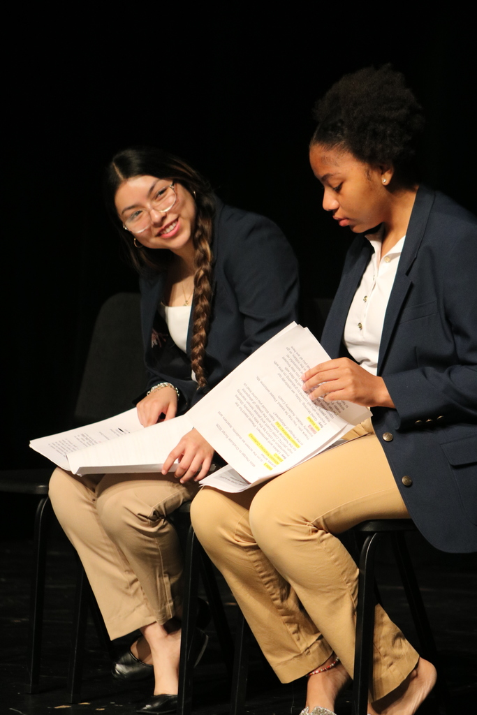 Two people sit side by side on stage, each holding printed documents with highlighted text. They wear navy blazers and light pants, and the background is dark.