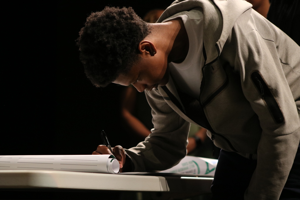 A student wearing a light gray hoodie leans over a table and signs a large printed pledge sheet. The background is dark, and the lighting focuses on the student and the paper.