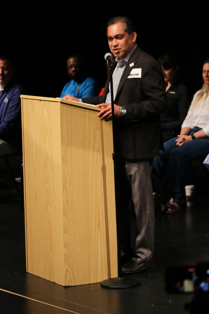 A person in a dark suit and checkered shirt speaks at a wooden podium. Several people sit behind them on stage, dressed in various colors. The setting appears to be a formal ceremony.