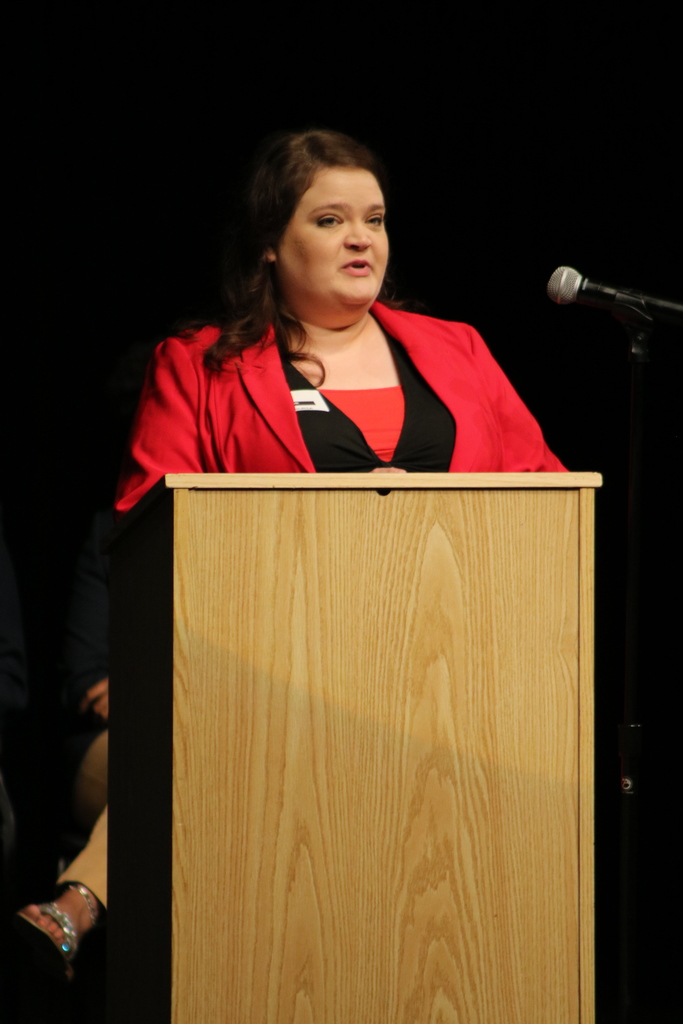A person wearing a red jacket stands behind a wooden podium speaking into a microphone. The background is dark, suggesting a stage setting.