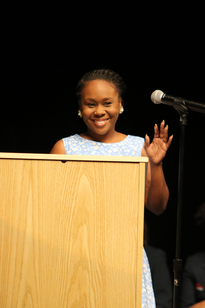 A person in a light blue dress with a white floral pattern stands at a podium, smiling and waving. A microphone is positioned in front of them, and the background is dark.