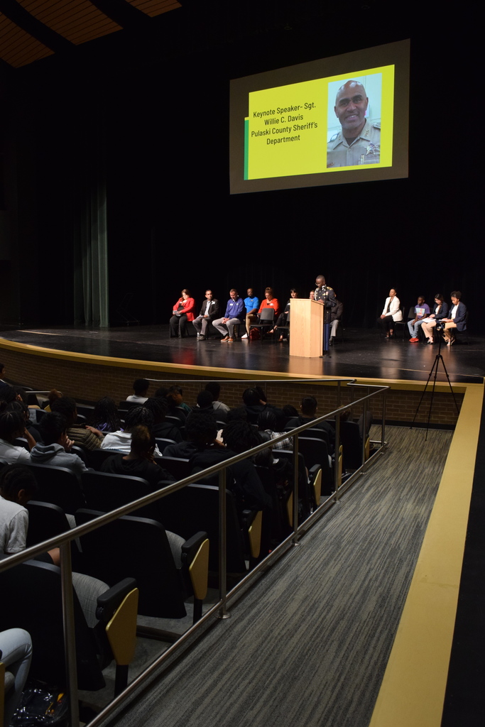 An auditorium audience faces a stage where several people are seated behind a podium. A large screen displays a slide reading “Keynote Speaker – Sgt. Willie Davis, Pulaski County Sheriff’s Department,” with his photo in uniform.