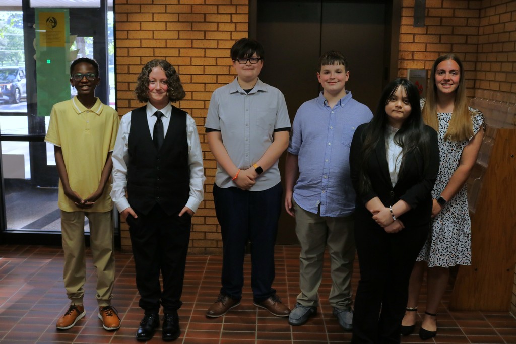 Five students and a teacher from the Sylvan Hills Middle School FBLA stand in a hallway near a brick wall and elevator, dressed in semi‑formal attire before entering the PCSSD school board meeting to present.