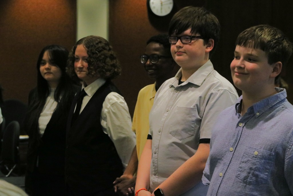 Five Sylvan Hills Middle School FBLA students stand in a line indoors, dressed in semi‑formal attire, waiting to present at the PCSSD school board meeting. Audience members are seated behind them.