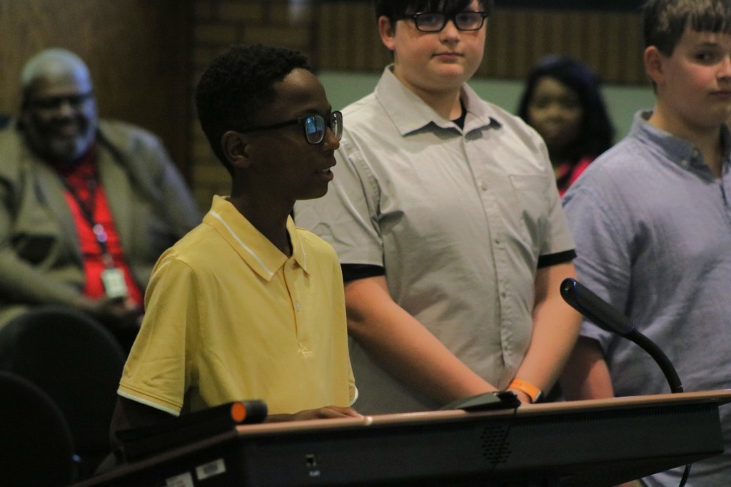 Three Sylvan Hills Middle School FBLA students stand at a podium during the PCSSD school board meeting. The student in front, wearing a yellow polo, speaks into the microphone while two others stand behind in support. Audience members are seated in the background.