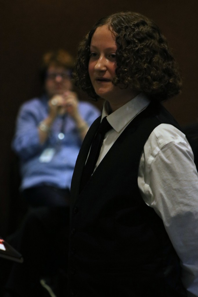 A Sylvan Hills Middle School FBLA student wearing a white shirt, black vest, and tie speaks during the PCSSD school board meeting. Another attendee sits in the background observing the presentation.