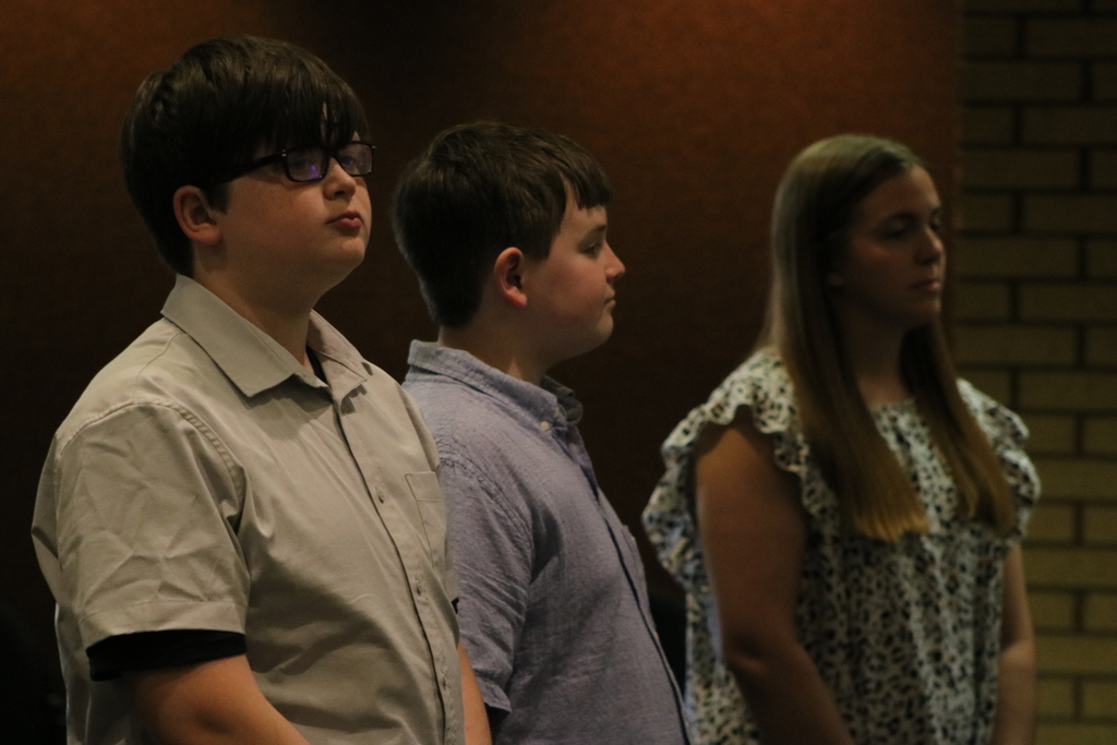 Three Sylvan Hills Middle School FBLA students stand side by side indoors, facing forward as they prepare to speak at the PCSSD school board meeting. They wear business‑casual clothing against a brown and brick wall backdrop.
