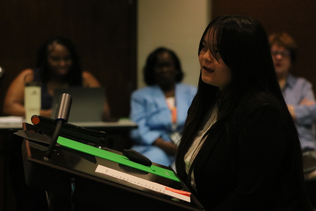 A Sylvan Hills Middle School FBLA student stands at a podium addressing the PCSSD school board. A green folder and printed notes rest on the podium. Others are in the background.