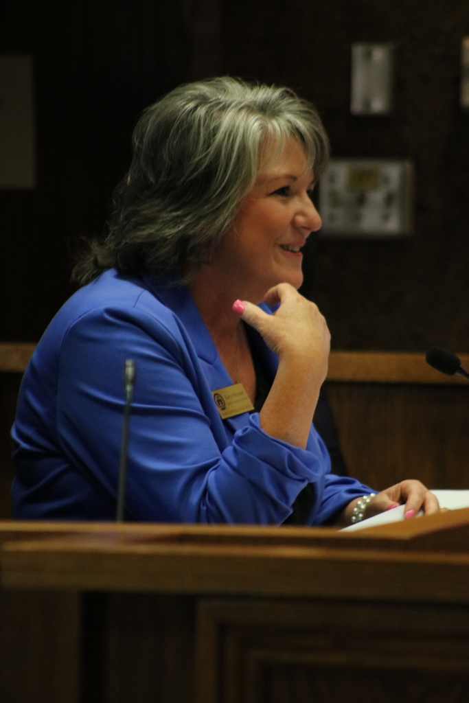 A woman in a bright blue blouse leans toward a podium while speaking at the PCSSD school board meeting. She holds a sheet of paper and appears engaged with the audience. A microphone and wooden paneling are visible around the podium.