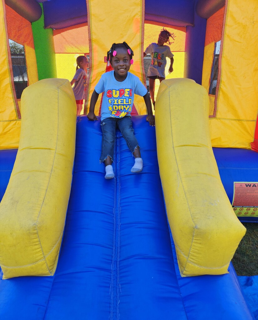 A child in a light blue shirt sits at the top of a blue inflatable slide inside a colorful bounce house.