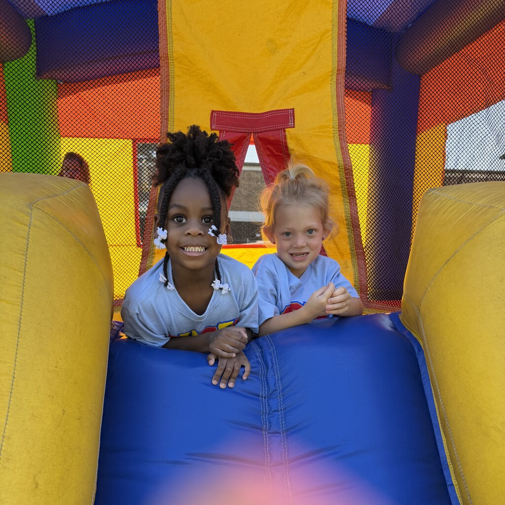 Two children in light blue shirts smile while lying on the blue floor of a colorful inflatable bounce house.
