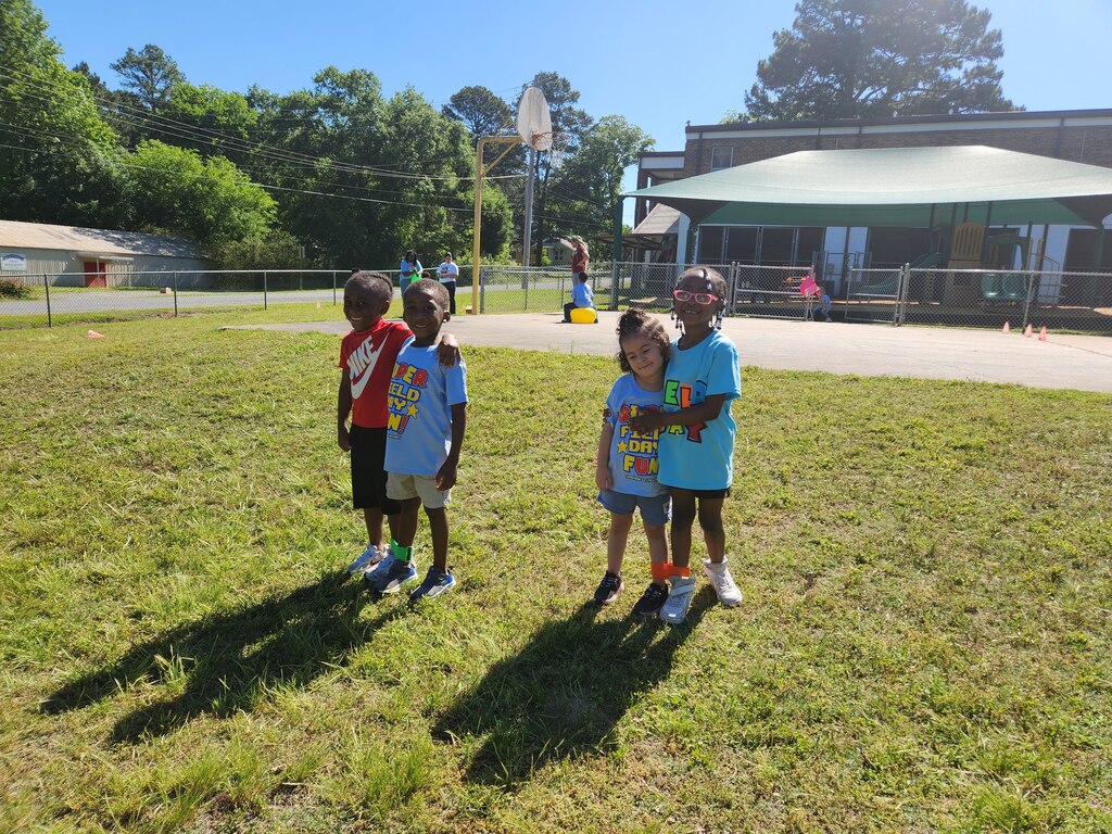 Four children stand in pairs with arms around each other on a grassy field near a basketball court during an outdoor activity.