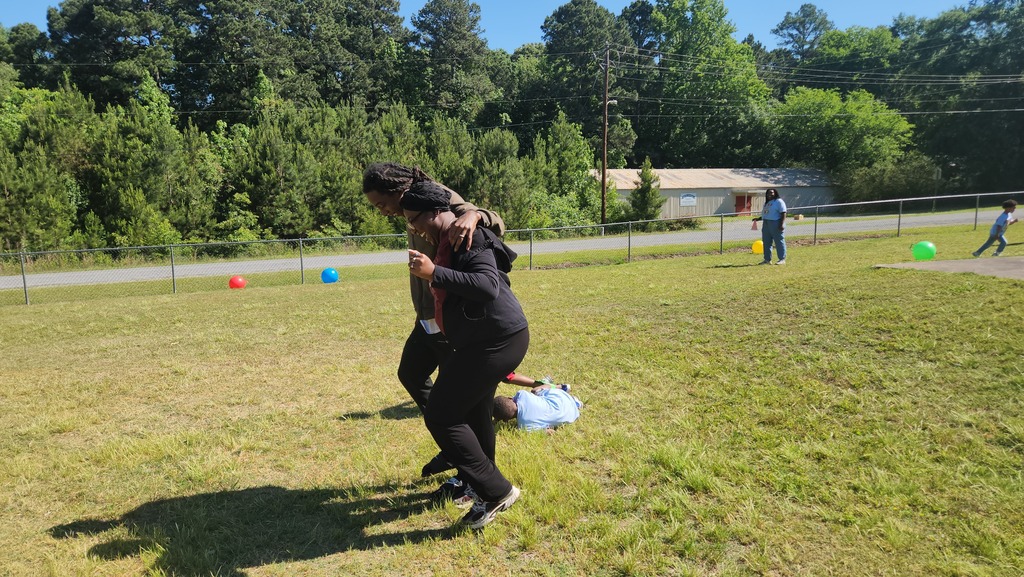 Two people with their legs tied together run across a grassy field during a three‑legged race, with colorful balls scattered in the background.
