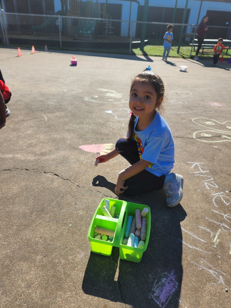 A child sits on pavement holding sidewalk chalk next to a container of chalk, with drawings and the word “Kinzie” visible on the ground.