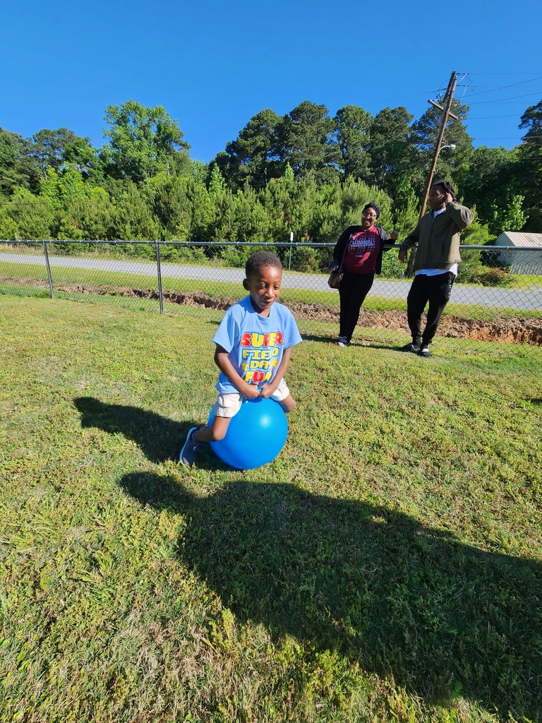 A child sits on a large blue ball in a grassy field while two adults stand near a fence in the background.