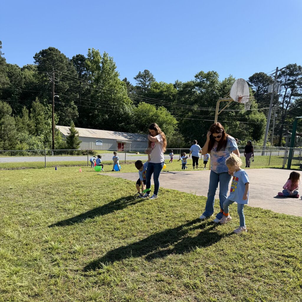 An adult holds a smiling child on a sunny day near a weathered basketball hoop at an outdoor play area.