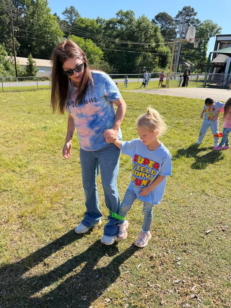 An adult and child with their legs tied together run hand‑in‑hand on a grassy field during a three‑legged race.