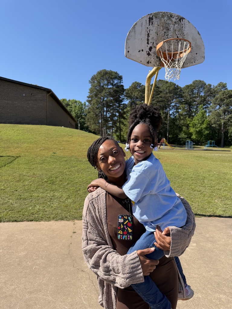 An adult holds a smiling child on a sunny day near a weathered basketball hoop at an outdoor play area.