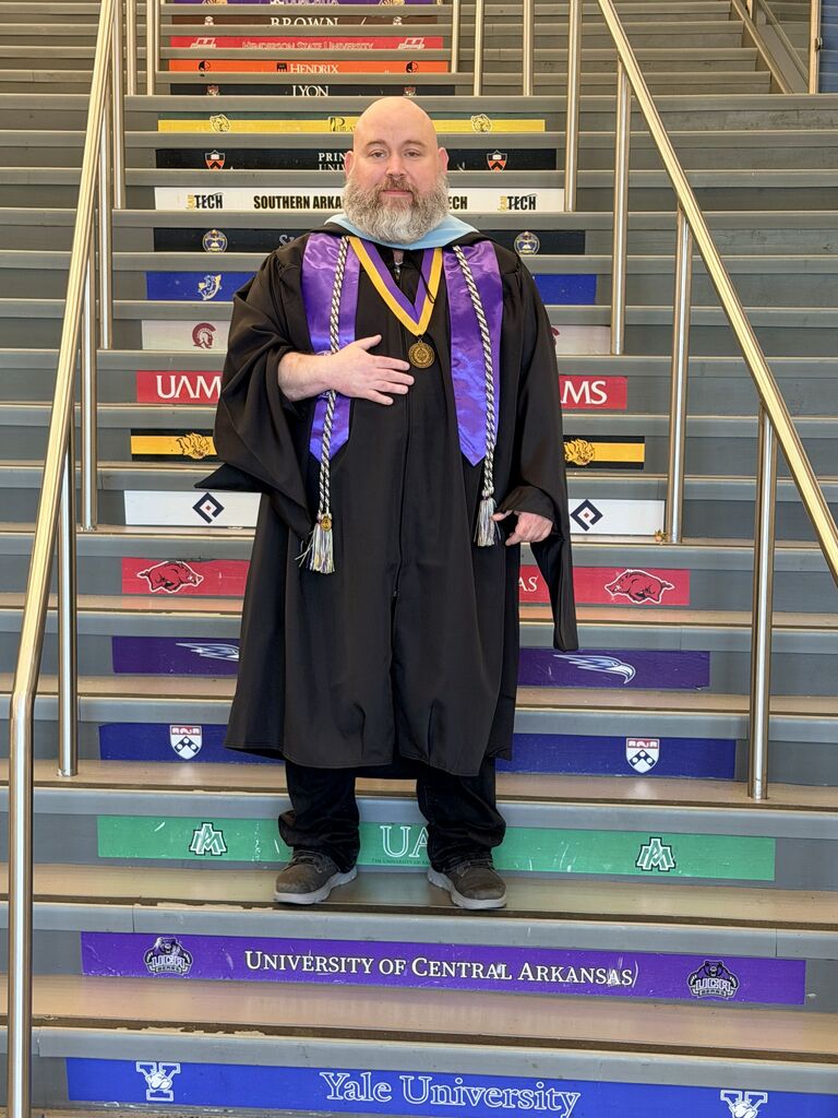 Chris Walker stands on a staircase decorated with colorful university names and logos while wearing a graduation gown with a purple stole.