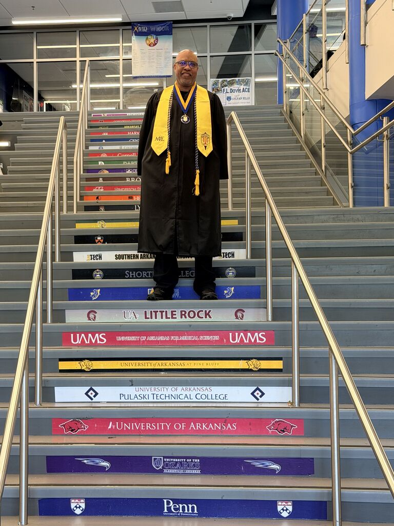 Officer Jamal Hockaday stands on a staircase featuring university names and logos while wearing a graduation gown with a yellow stole.