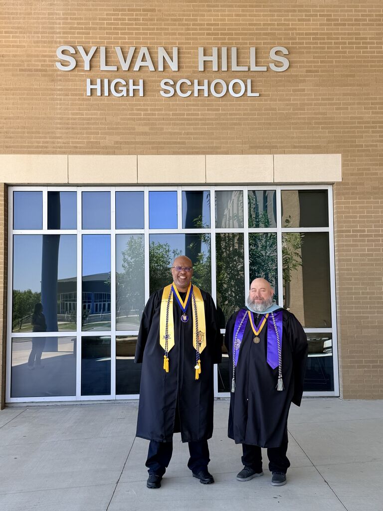 Chris Walker and Officer Jamal Hockaday stand side by side in graduation gowns and stoles in front of the Sylvan Hills High School building.