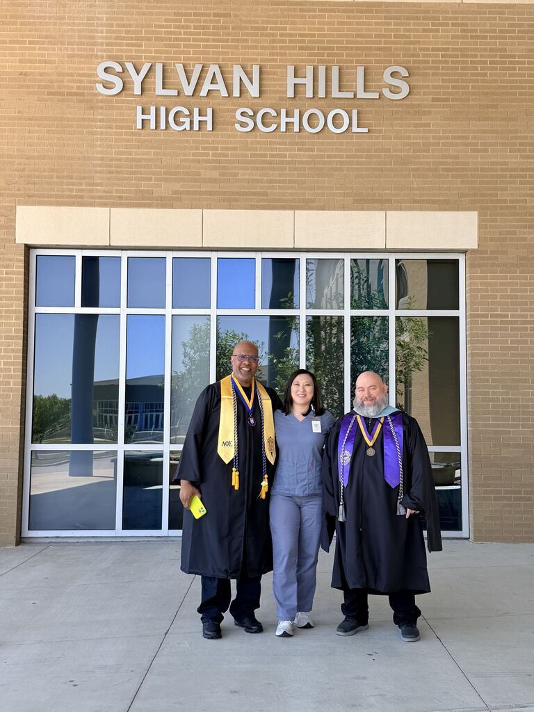 Chris Walker, a person in scrubs, and Officer Jamal Hockaday stand together in front of the Sylvan Hills High School building.