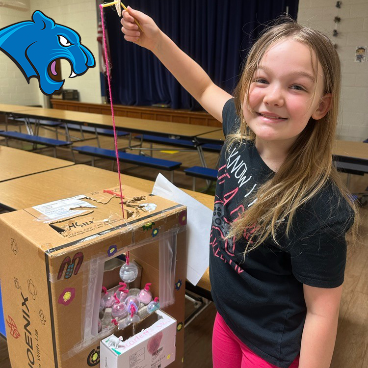 A child holds a homemade cardboard claw machine filled with toy capsules in a school cafeteria.