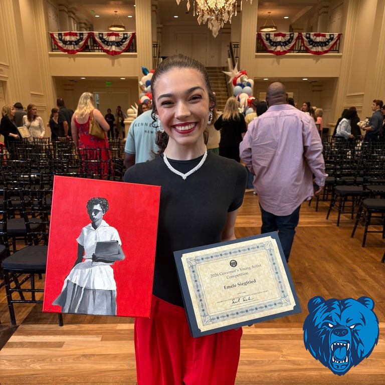 A student stands in a decorated hall holding a painting and a certificate from the 2026 Governor’s Young Artist Competition.