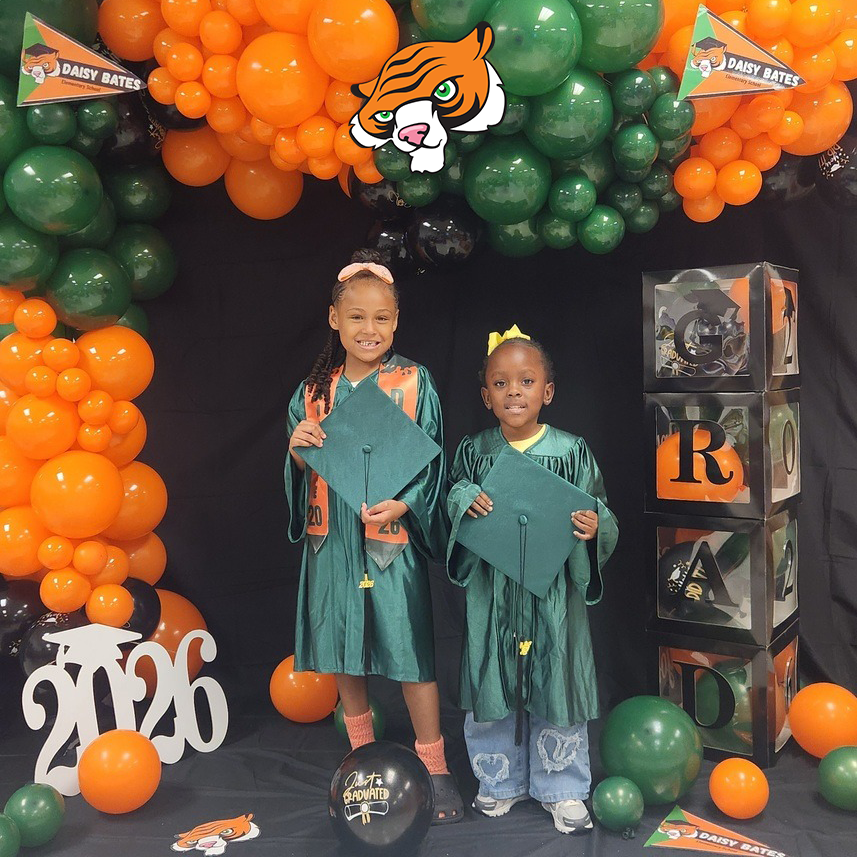 Two children in green graduation gowns hold their caps while standing in front of a balloon backdrop with a tiger mascot and “GRAD” boxes.
