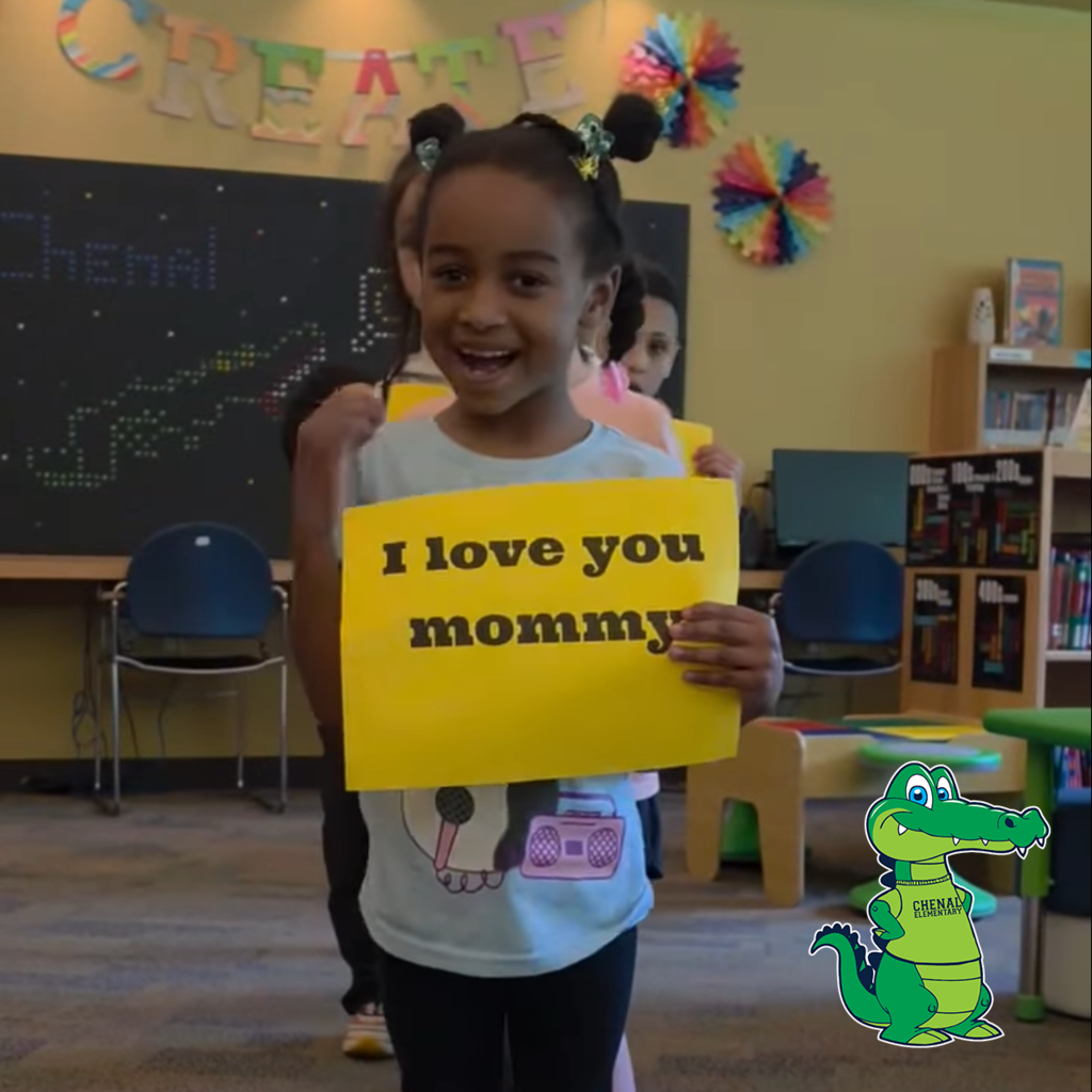 A child holds a yellow sign reading “I love you mommy” while other children hold similar signs in a classroom.