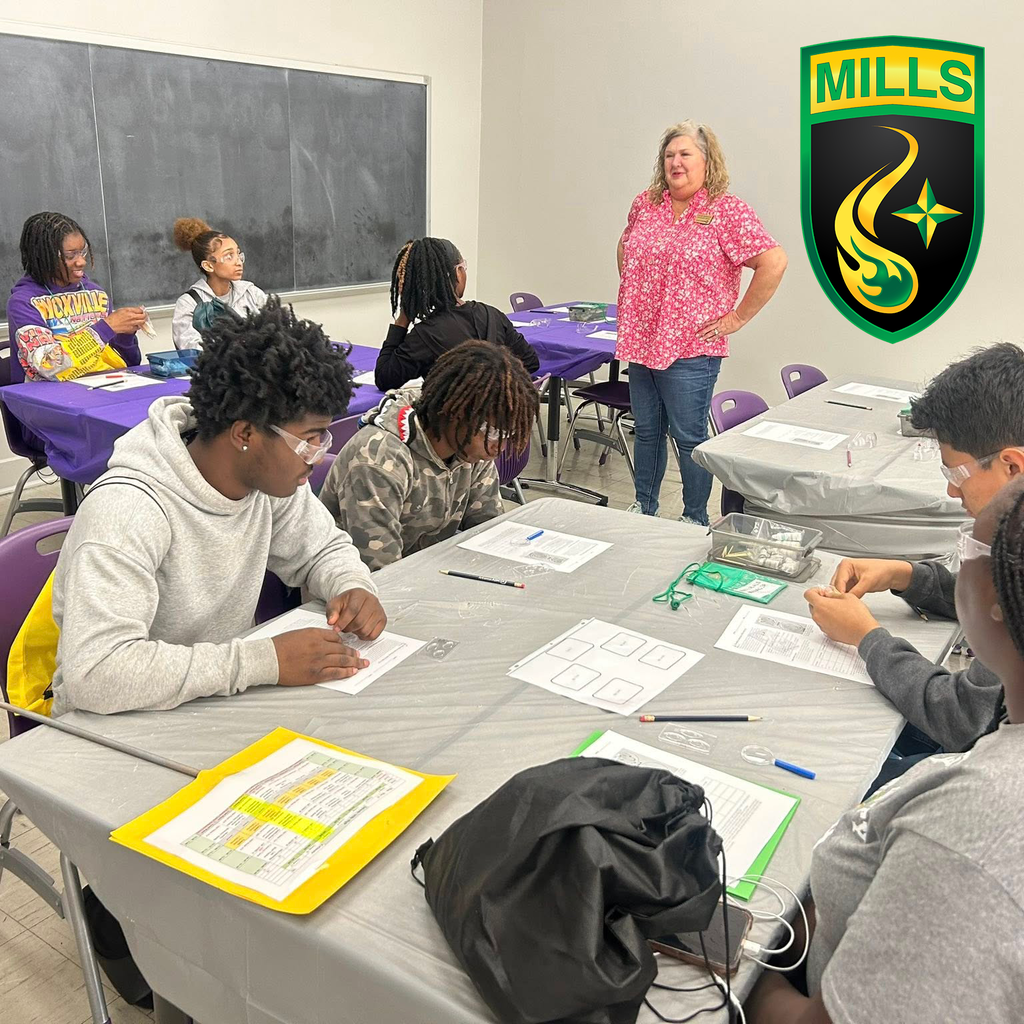 Students sit at tables working on worksheets while a teacher leads a lesson at the front of the classroom.