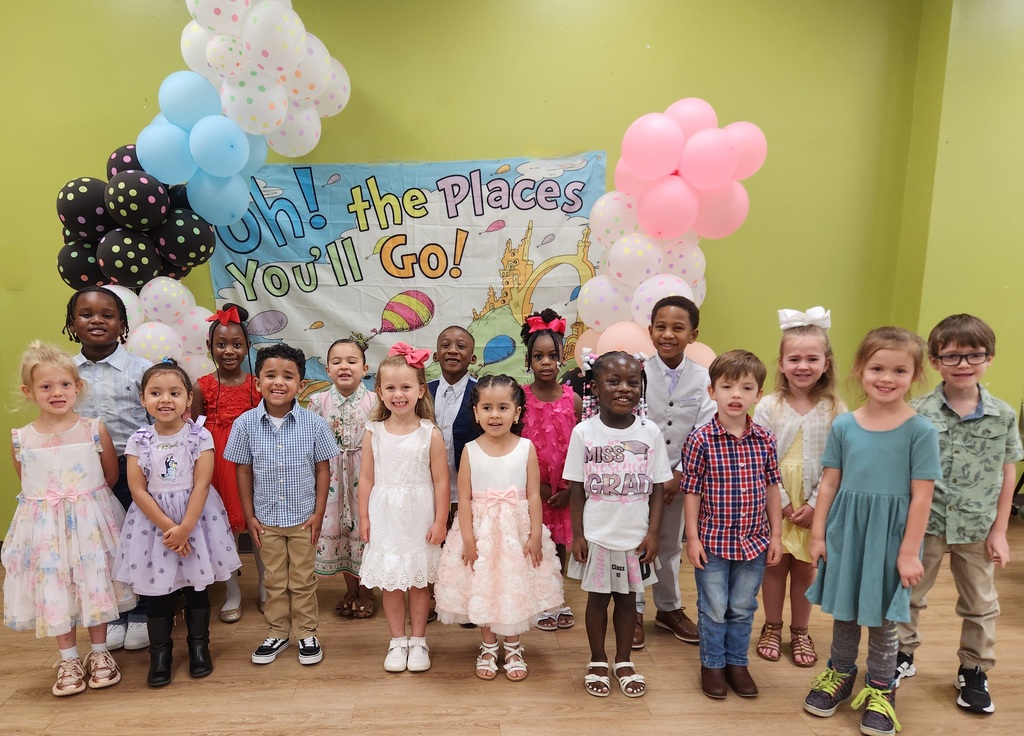 Young children stand in front of a colorful “Oh! the Places You’ll Go!” graduation backdrop surrounded by pastel balloons.