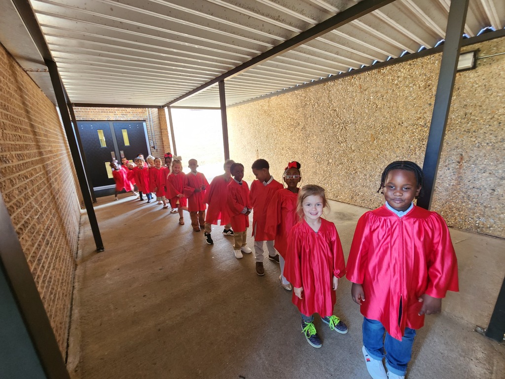 A group of Pre‑K students wearing red graduation gowns walk in a line down a covered walkway toward a set of double doors.
