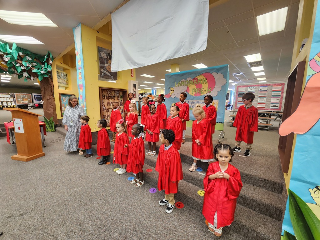 Pre‑K students in red graduation gowns stand in rows in front of a “Landmark Pre‑K” backdrop while an adult stands beside a podium labeled “Cardinals.”