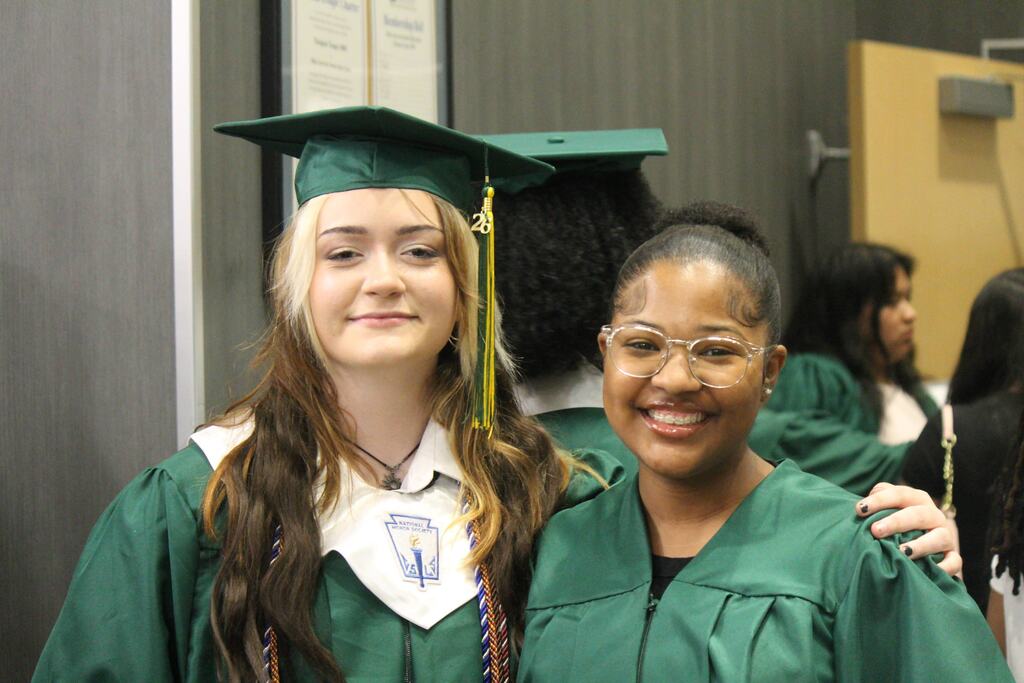 Two graduates in green caps and gowns stand together smiling; one wears a National Honor Society collar and cords, and the other wears clear glasses. Other graduates are visible behind them.