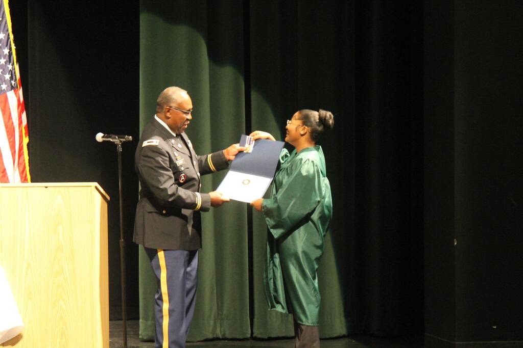 A person in a military uniform presents a certificate to a graduate in a green gown on a stage with a podium, microphone stand, and U.S. flag.