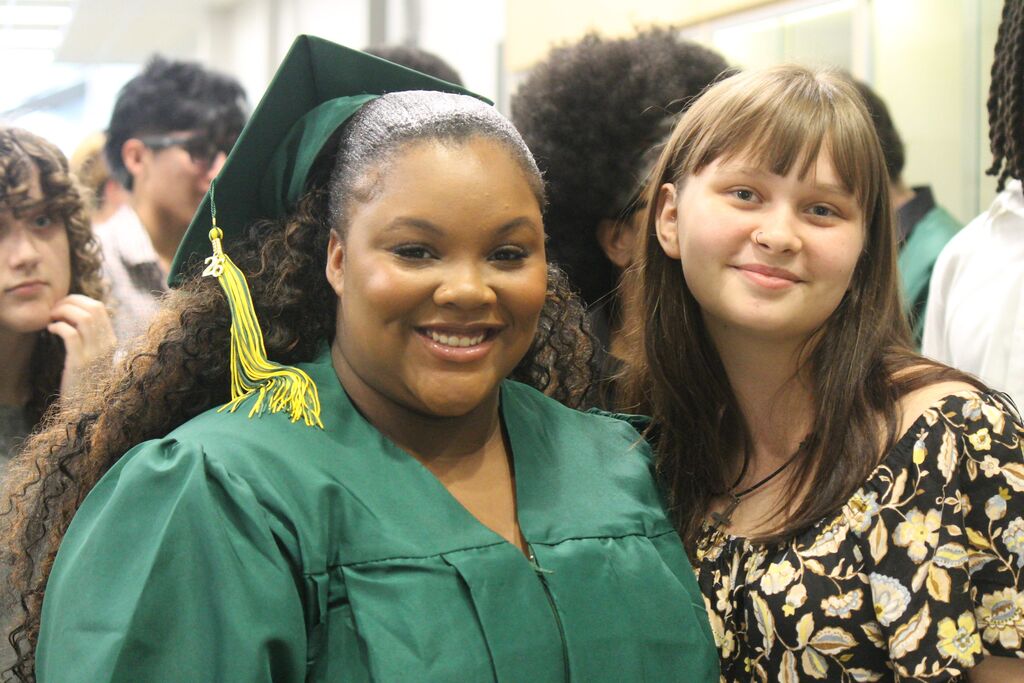 A graduate in a green cap and gown with a “26” tassel stands closely with another person in a floral blouse in a crowded indoor space with other graduates in the background.