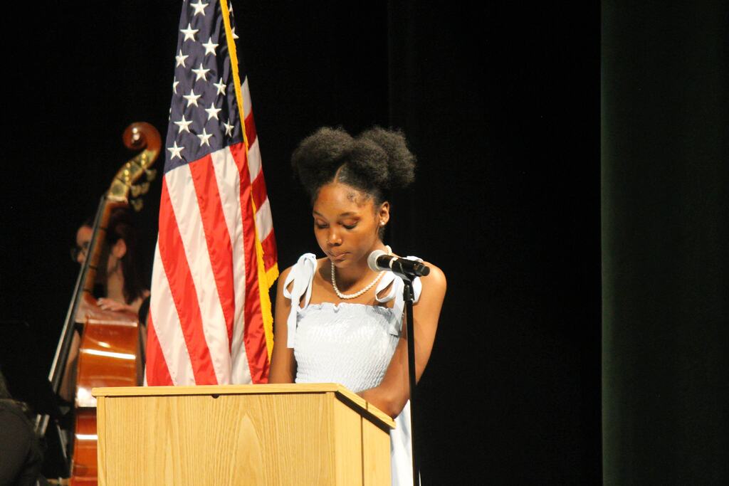Two people wearing formal attire with ribbons stand on a stage; one speaks at a podium while the other holds an item in both hands. A U.S. flag and a table with a white cloth are visible.