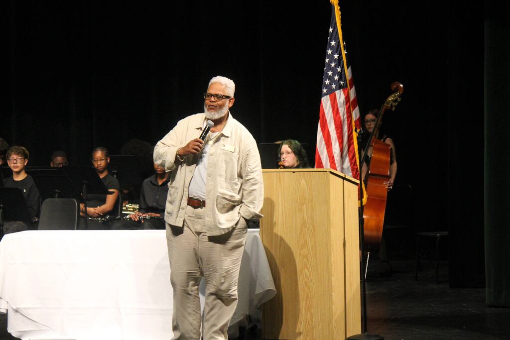 A person holding a microphone speaks onstage near a podium with a U.S. flag, while musicians with instruments sit behind them.