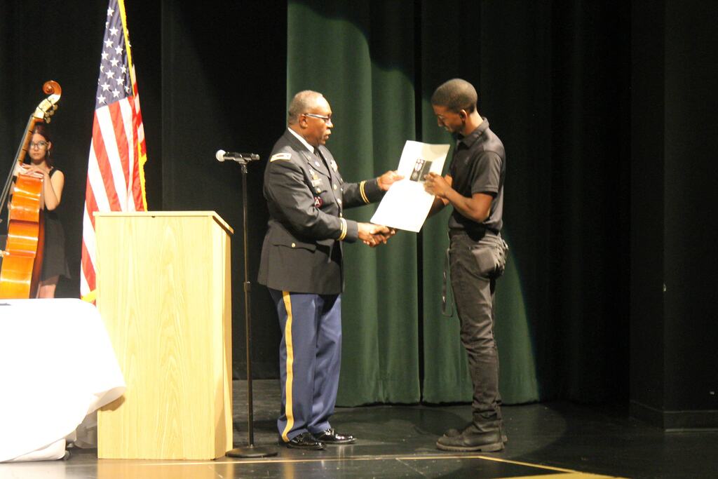 A person in a military uniform hands a certificate to someone dressed in black on a stage with a podium, U.S. flag, and a musician standing near a large string instrument.