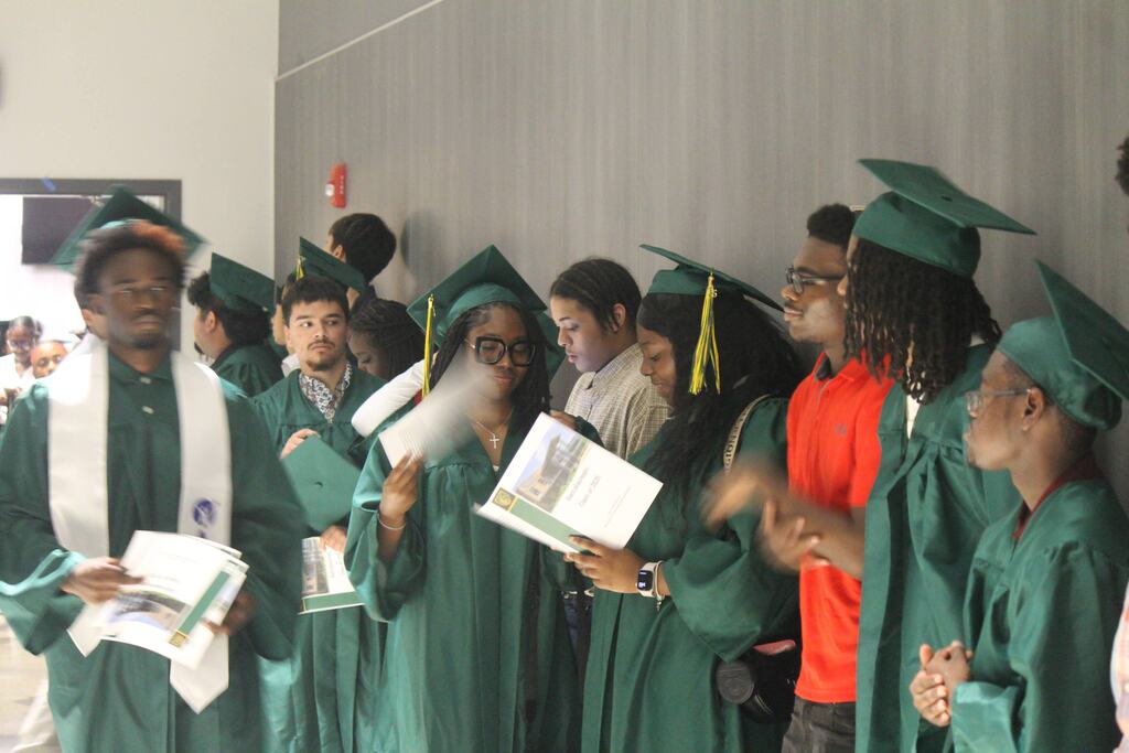 A group of graduates in green caps and gowns stand in a hallway holding programs, with other people and a gray wall in the background.